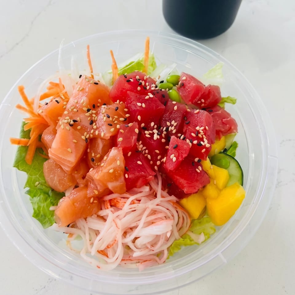 Colorful poke bowl with salmon, tuna, mango, cucumber, and crab sticks in a clear plastic container.